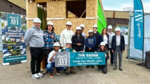 A group of people, mostly wearing hard hats, stand smiling in front of a partially constructed house. They hold a banner that says, THE SASHA and Proudly built with M.E. Lazerte High School Students.
