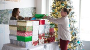 Two people stack colorful gift boxes on a table beside a decorated Christmas tree with white and blue ornaments in a brightly lit room.