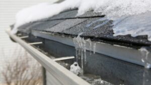 A close-up of a house roof with melting snow and icicles hanging from the edge, above a metal gutter partially filled with ice.