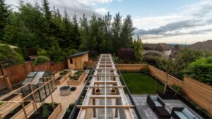 A modern backyard features raised garden beds, a wooden pergola, a greenhouse, a small shed, gravel paths, a fire pit, and a grassy lawn, all enclosed by wooden fencing with trees in the background.