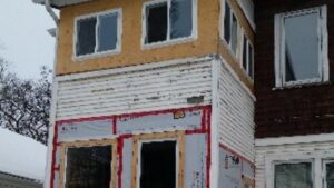 A house under renovation with new white siding and exposed plywood near the windows, along with construction materials and tape visible on the lower section.
