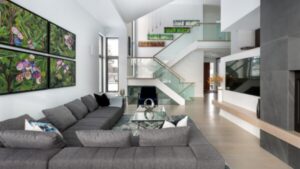 Modern living room with a gray sectional sofa, glass coffee table, large wall art, and a staircase with glass railings leading to an upper floor. Light wood flooring and a built-in TV are visible.
