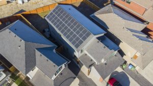 Aerial view of a house with solar panels on its roof, surrounded by neighboring houses and wooden fences, on a sunny day.