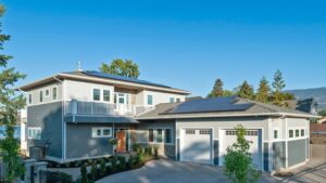 Modern two-story gray house with white trim, solar panels on the roof, three-car garage, large driveway, and green trees around, set against a clear blue sky.