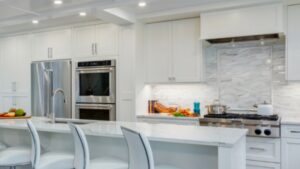 Modern white kitchen with marble countertops, stainless steel appliances, a stovetop with pots, a double oven, and four white barstools at a large island. Fresh vegetables and kitchen items are on the counters.