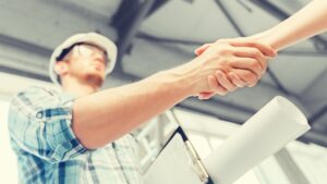 A construction worker wearing a hard hat and holding blueprints shakes hands with another person, symbolizing agreement or partnership at a worksite.