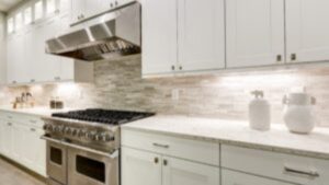 Modern kitchen with white cabinets, a stainless steel gas stove, a range hood, light stone backsplash, and two white jars on a marble countertop. The space is well-lit with under-cabinet lighting.