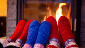 Four pairs of feet wearing colorful, patterned socks rest in front of a lit fireplace, suggesting a cozy and warm indoor setting.