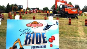 A sign reading “Take a Ride for Mackids” is in front of a grassy area with construction vehicles, orange safety barrels, and people gathered under a cloudy sky.