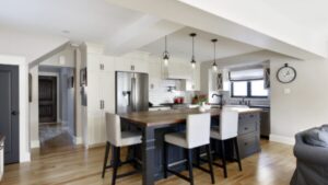Modern kitchen with a central island, three chairs, stainless steel appliances, white cabinets, pendant lights, a clock on the wall, and wooden floors. Bright and clean design with natural light.