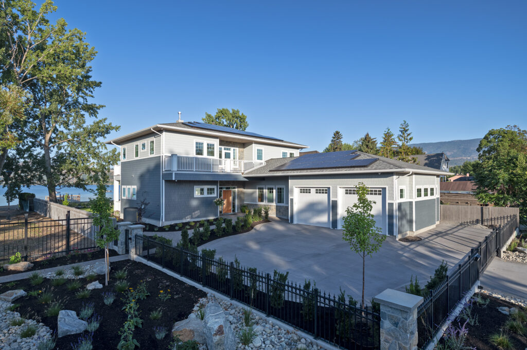 Modern two-story house with gray siding, large windows, a three-car garage, solar panels on the roof, landscaped front yard, black fence, and trees, set against a clear blue sky and distant mountains.