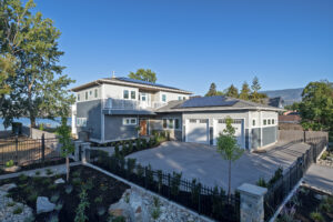 Modern two-story house with gray siding, large windows, a three-car garage, solar panels on the roof, landscaped front yard, black fence, and trees, set against a clear blue sky and distant mountains.