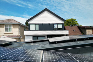 Modern house with white siding and black accents, set behind multiple solar panels installed on nearby rooftops, under a blue sky with scattered clouds—an ideal example of Net Zero Homes during power outages.