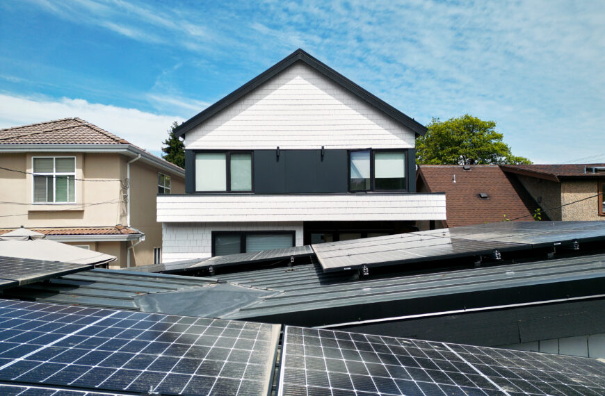 Modern house with white siding and black accents, set behind multiple solar panels installed on nearby rooftops, under a blue sky with scattered clouds—an ideal example of Net Zero Homes during power outages.