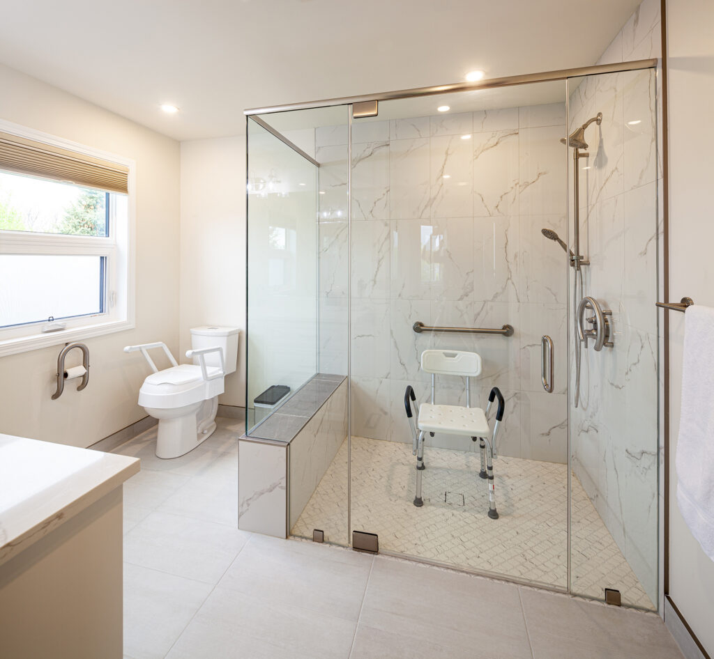 Accessible bathroom with a glass-enclosed walk-in shower containing a mounted shower seat and grab bar, alongside a toilet with removable armrests.