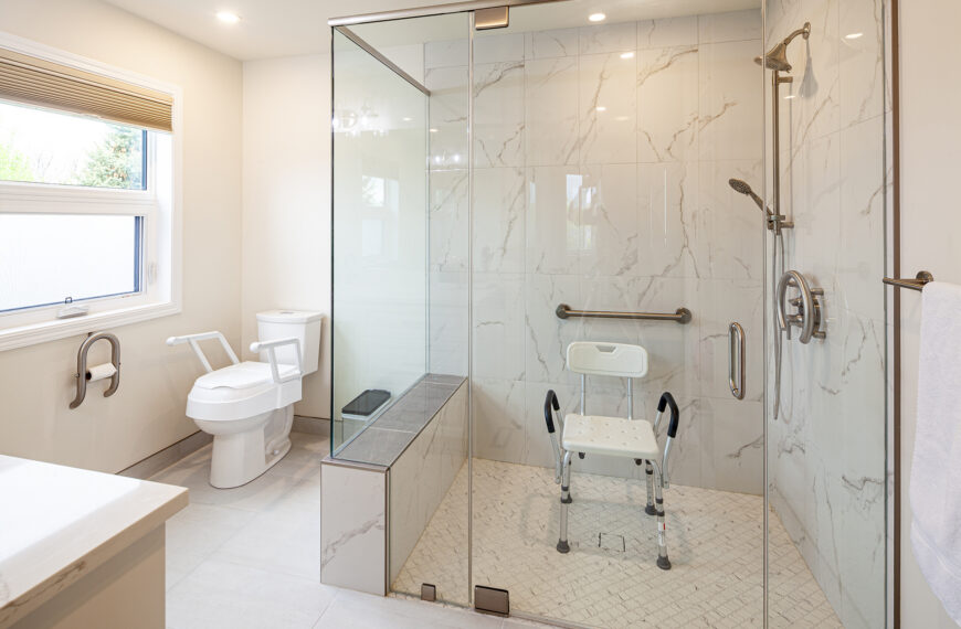 Accessible bathroom with a glass-enclosed walk-in shower containing a mounted shower seat and grab bar, alongside a toilet with removable armrests.