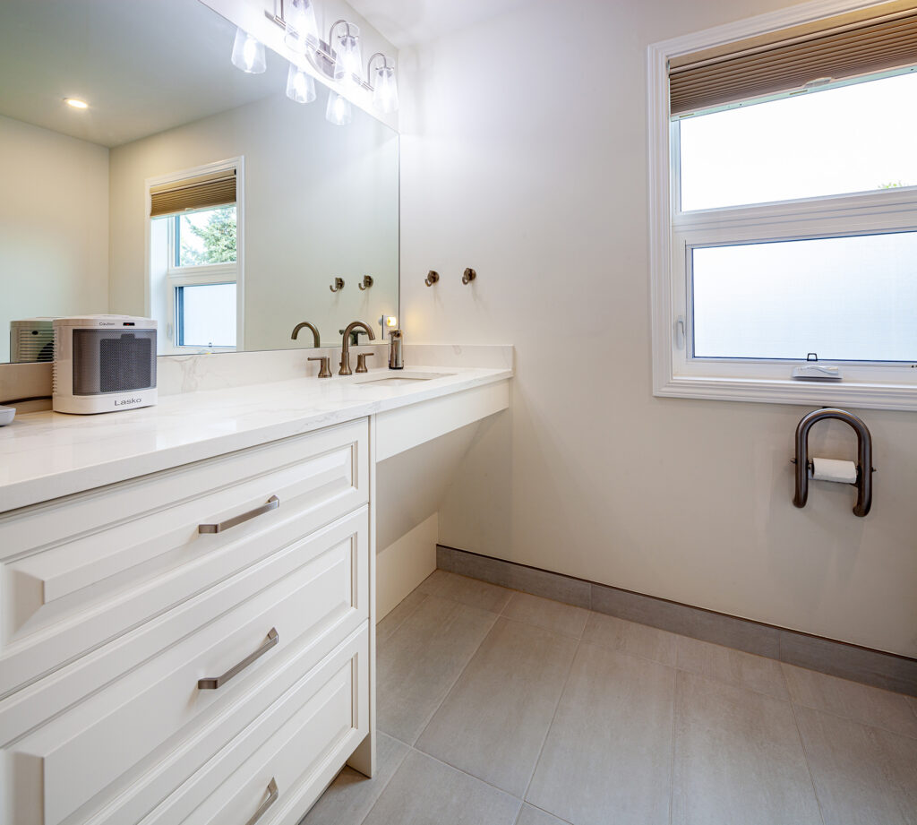 A bathroom vanity with a white countertop, a Lasko heater on the left, and a window with a toilet paper holder mounted below on the right, showcasing an aging in place renovation.