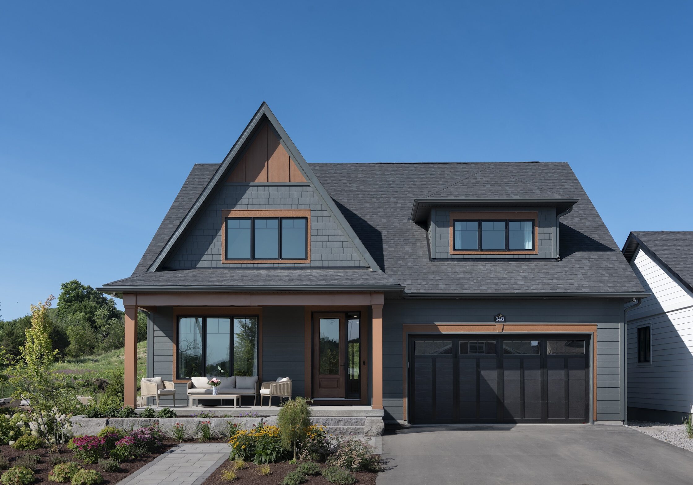 A modern two-story net zero home with dark gray siding, a steep gabled roof, large windows, a double garage, and a front porch with outdoor seating, surrounded by landscaped bushes and plants under a clear blue sky.