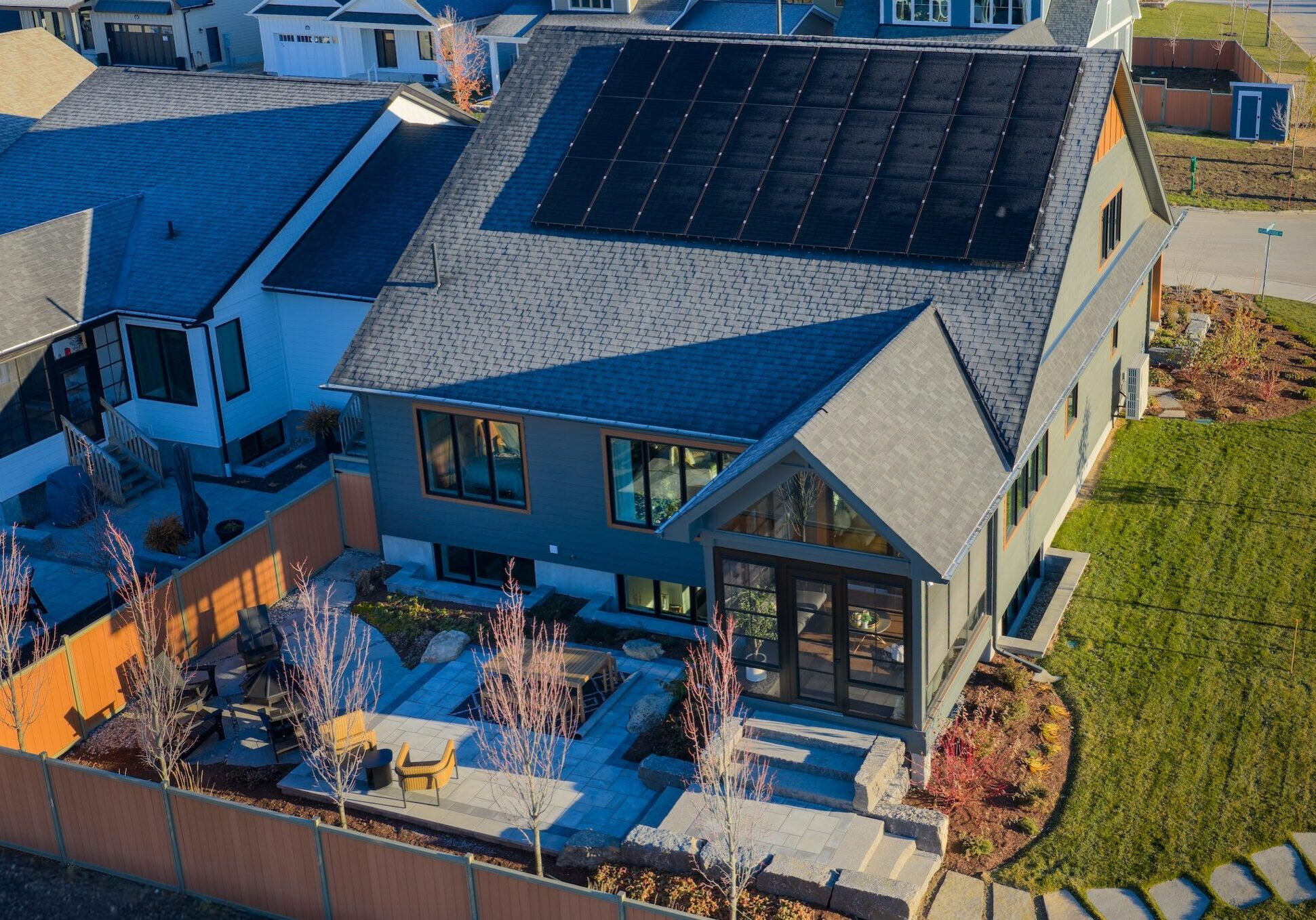 A modern net zero house with solar panels on the roof, a landscaped backyard with stone paths and patio seating, surrounded by a wooden fence in a suburban neighborhood.